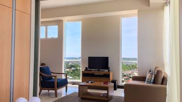 Quarto de hotel com varanda, televisão, sofá, cadeira e toalhas sobre uma mesa.
