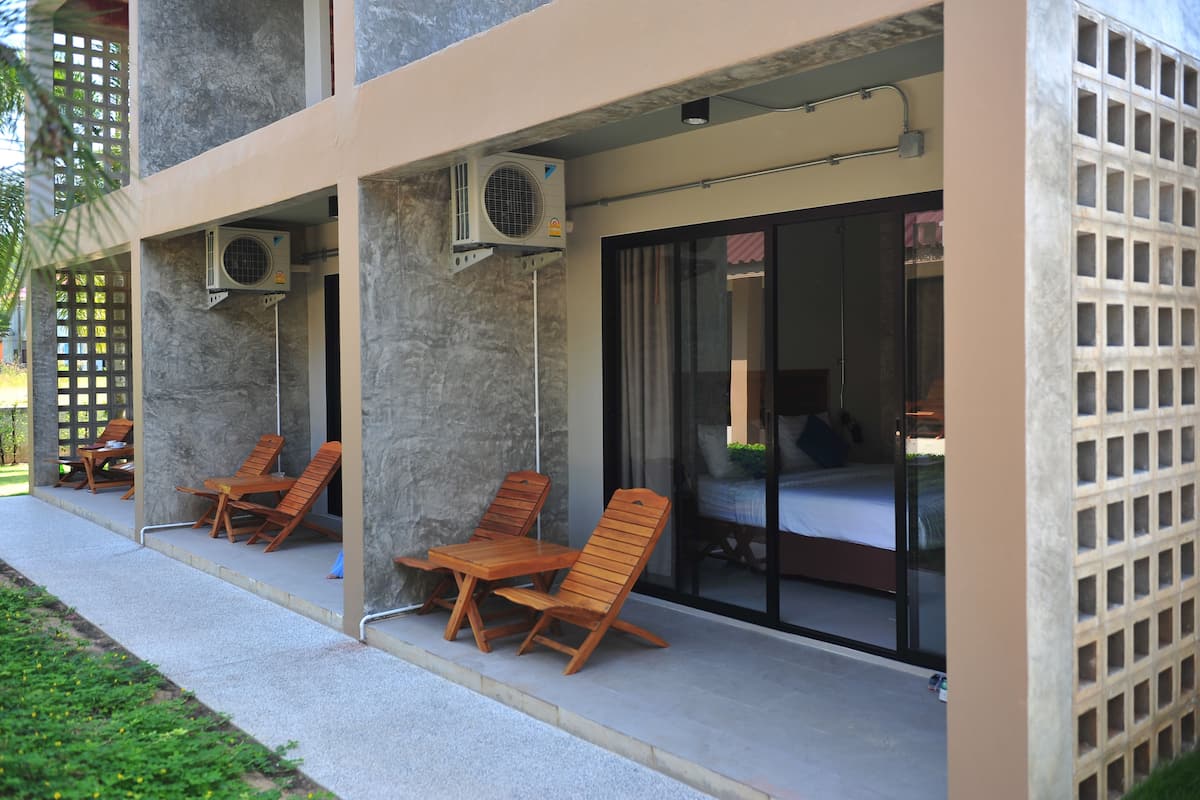 Uma área de estar coberta ao ar livre, com cadeiras e mesas de madeira, aparelhos de ar condicionado e uma porta de vidro que dá para um espaço interno.