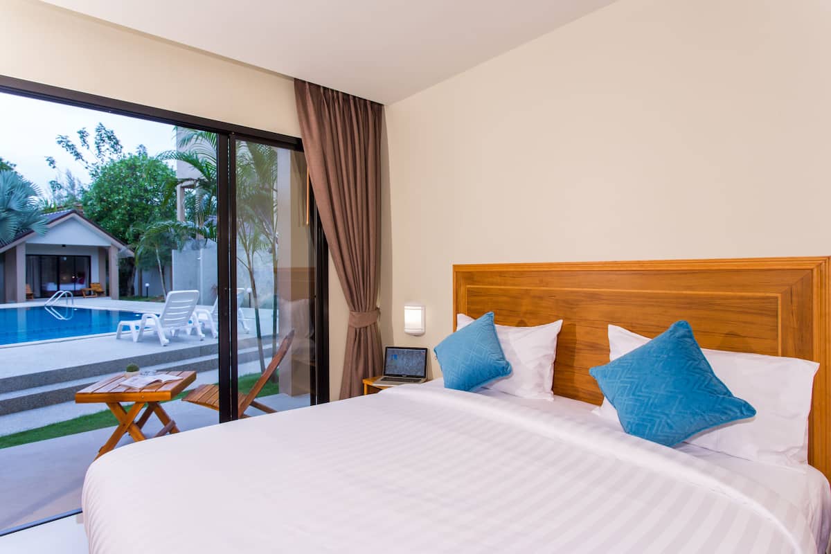 Quarto de hotel com uma cama grande, duas almofadas, um laptop sobre uma mesinha de cabeceira e vista para uma piscina e área externa com assentos.