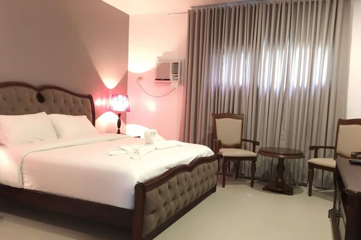 A hotel room with a large bed, a bedside lamp, a chair, a small table, and a window with curtains.