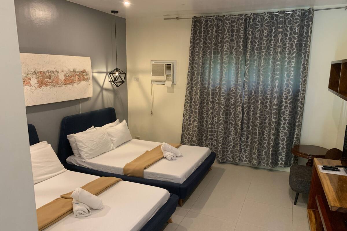 A hotel room with two beds, a desk, a chair, a TV, a patterned curtain, and a wall-mounted air conditioner.