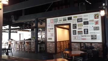 Interior de un restaurante con barra, mesas y sillas de madera. Se ven estantes con botellas y un panel con varios anuncios.