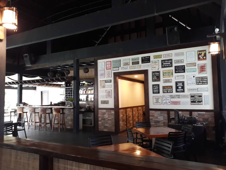 Interior de un restaurante con barra, mesas y sillas de madera. Se ven estantes con botellas y un panel con varios anuncios.