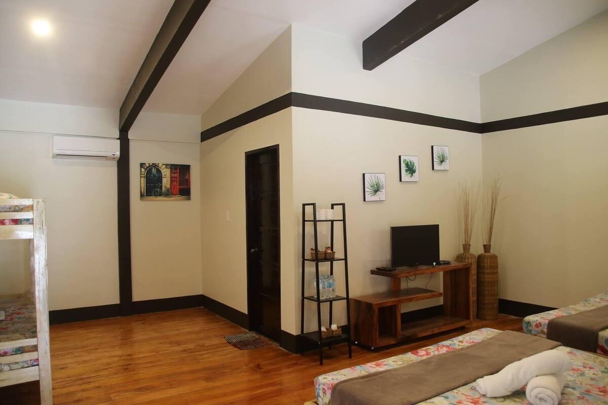 A hotel room with a wooden floor, a bed with a floral bedspread, a TV on a wooden stand, a black shelf with decorative items, and a door leading to another room.
