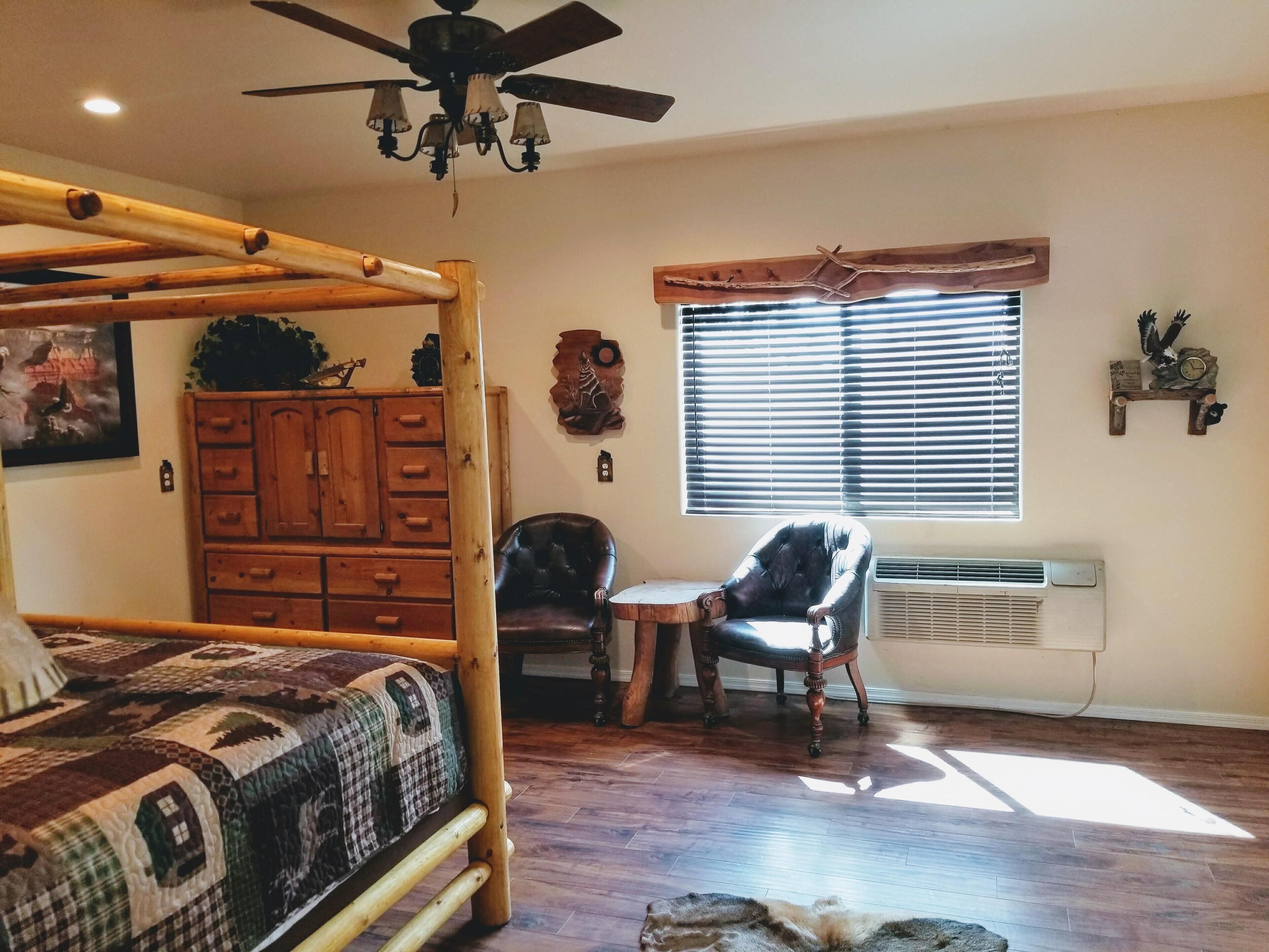 Une chambre avec un lit en bois, une chaise, une petite table, une fenêtre avec des stores, un ventilateur de plafond et un radiateur fixé au mur.