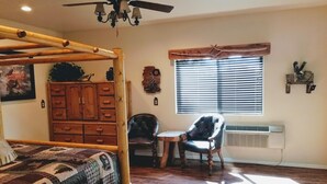 A bedroom with a wooden bed, a chair, a small table, a window with blinds, a ceiling fan, and a wall-mounted heater. - Mountain Views and Great Fire Pit (Star Valley)