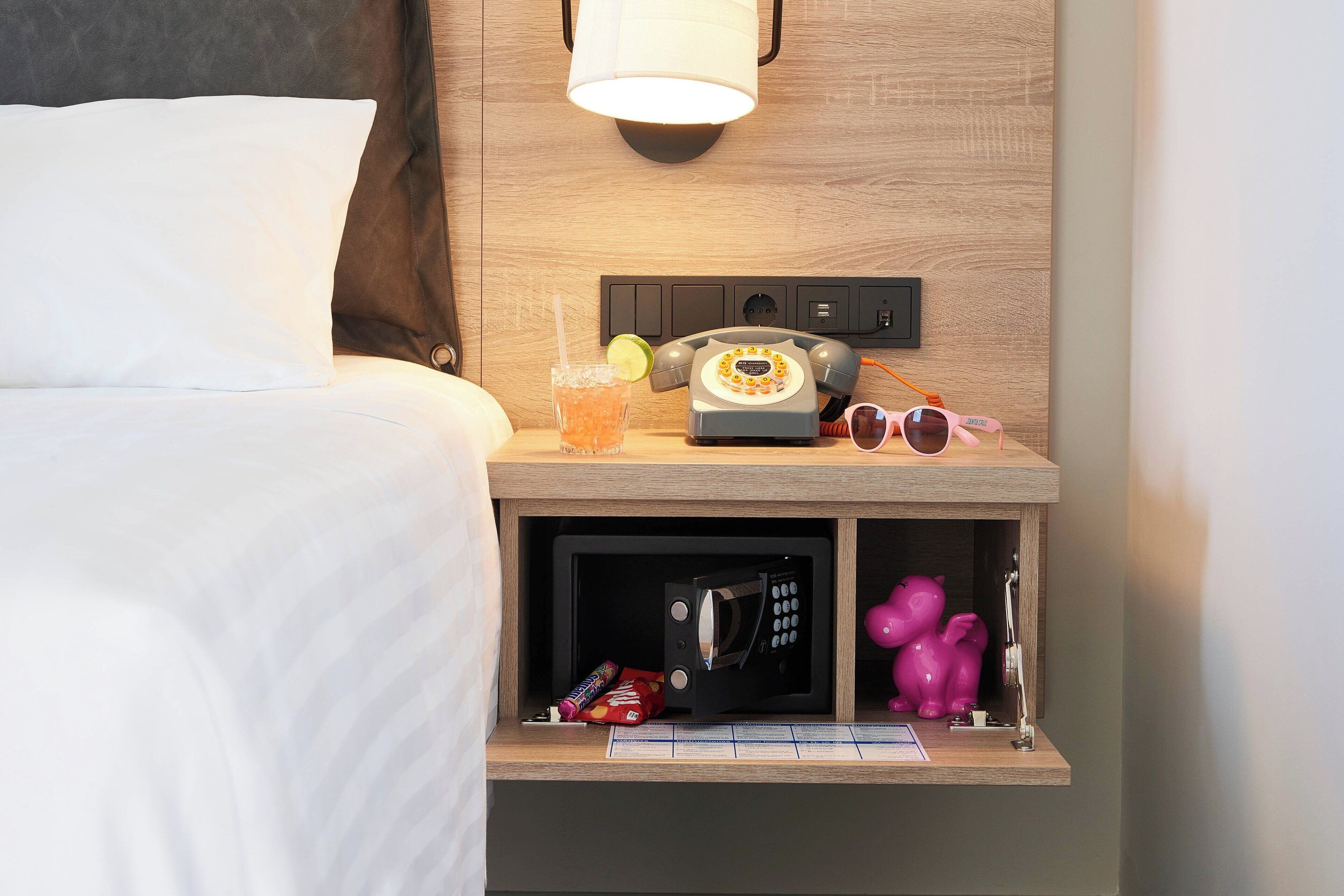 Un lit bien fait, avec un oreiller, une lampe de chevet, un téléphone, un verre avec une paille et une tranche de citron vert, un four à micro-ondes, un jouet rose et un paquet de collations sur la table de chevet.