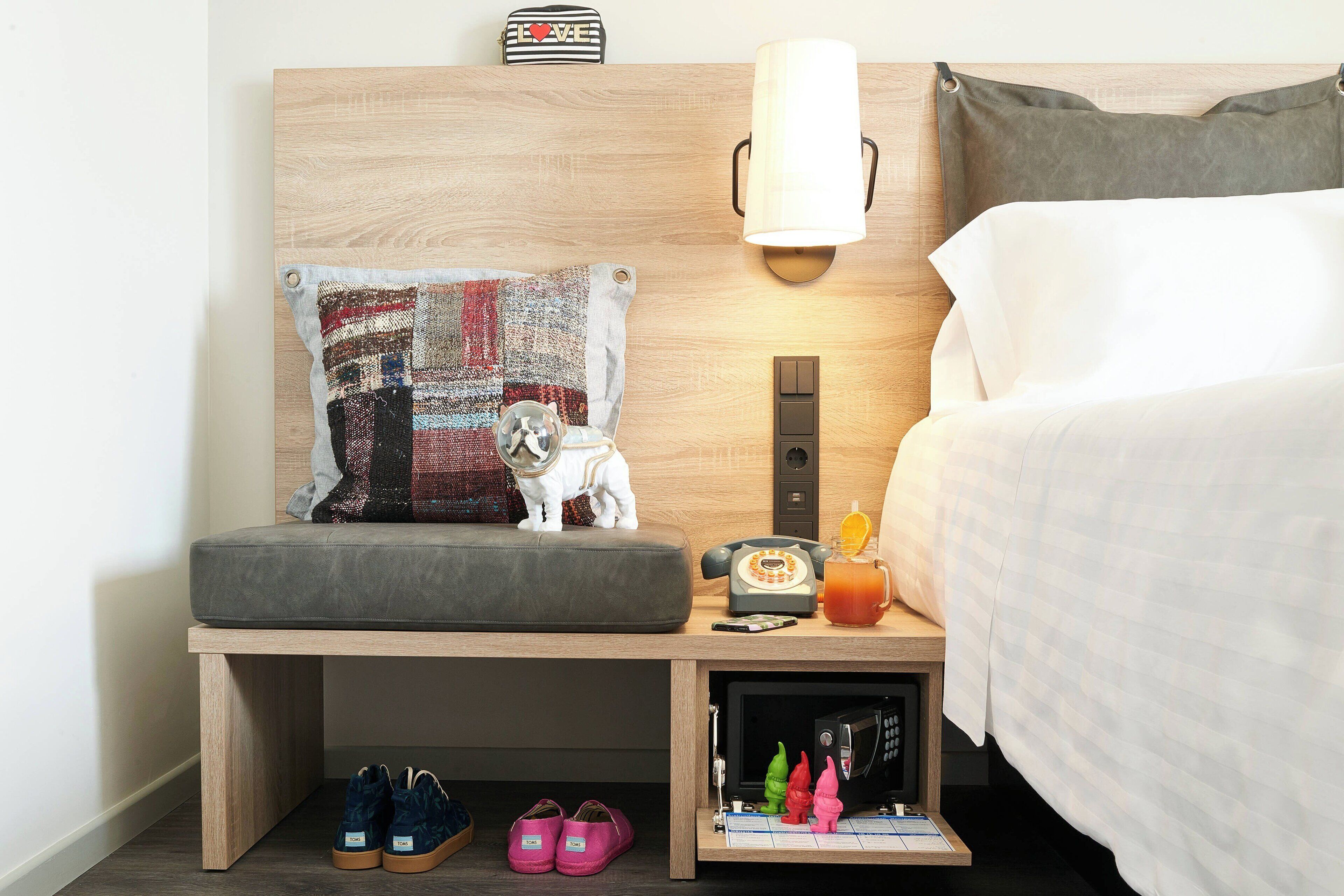 A neatly arranged hotel room with a bed, a bedside table with a lamp, a small table with a phone and a drink, and a shelf with shoes and a microwave.