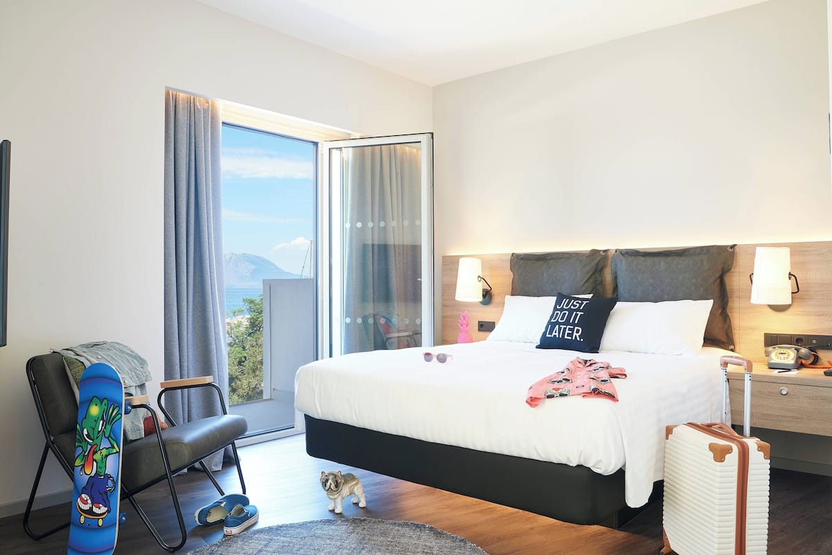 A hotel room with a bed, a chair, a suitcase, and a view of the outdoors.