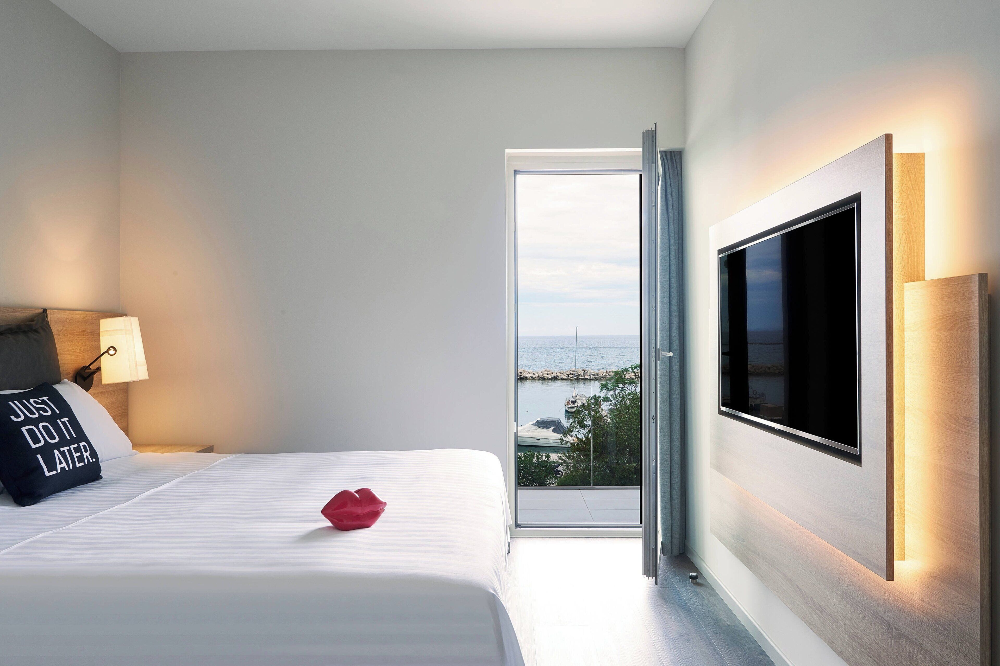 Une chambre d’hôtel moderne, équipée d’un lit, d’un téléviseur fixé au mur et offrant une vue sur la mer par une porte ouverte.