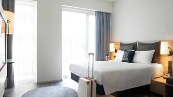 Una habitación de hotel con una cama, una maleta, un tapete redondo y una mesita de noche con una lámpara.