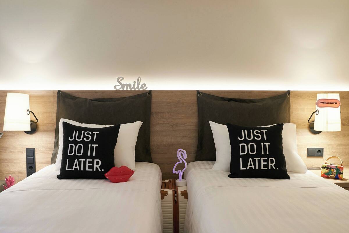 A hotel room with two beds, each with a pillow that reads "JUST DO IT LATER." A wall-mounted lamp and a sign that says "Smile" are visible.
