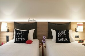 A hotel room with two beds, each with a pillow that reads "JUST DO IT LATER." A wall-mounted lamp and a sign that says "Smile" are visible.