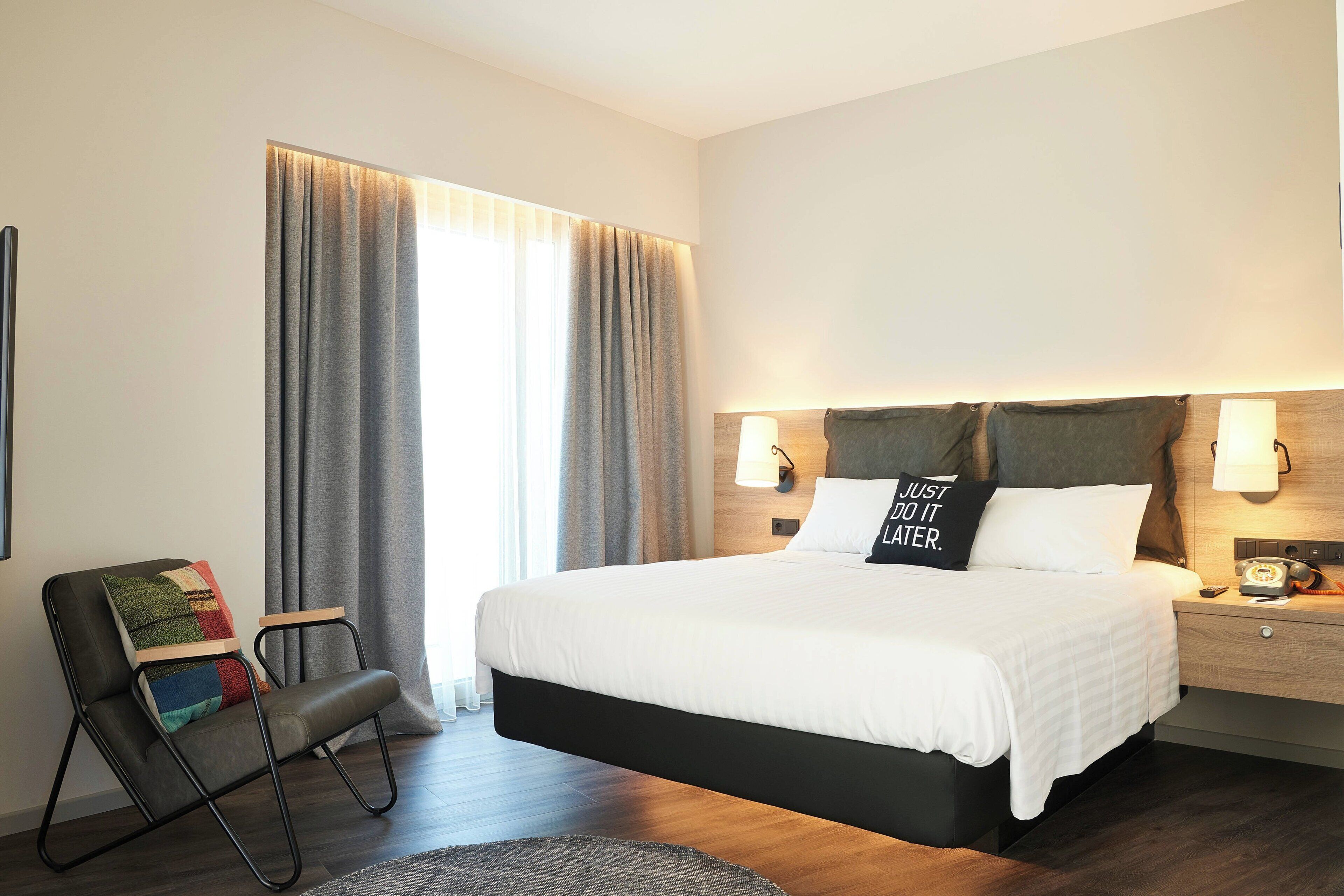 A modern hotel room with a large bed, a chair with a colorful blanket, a nightstand with a lamp, and a view of the outdoors through curtains.