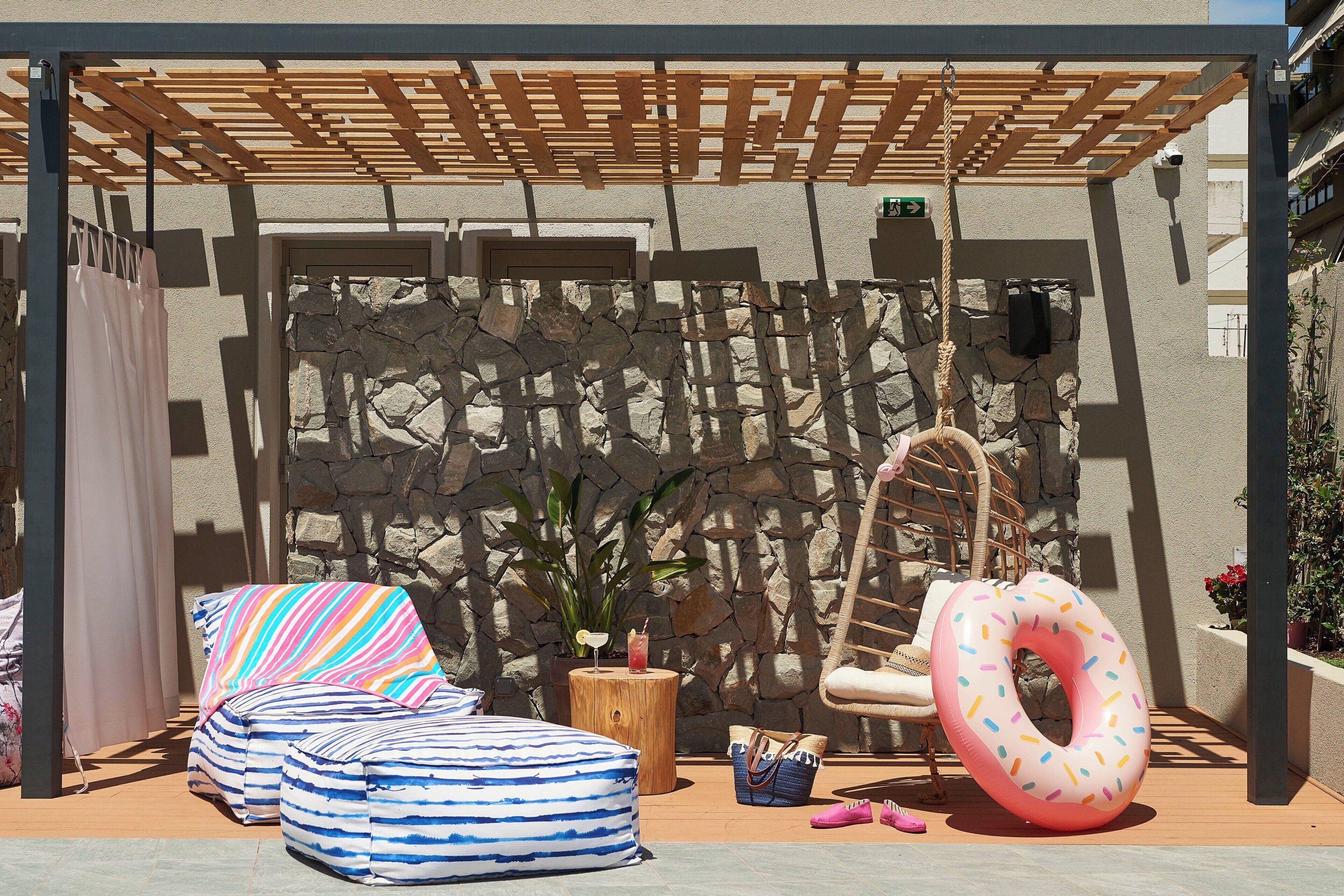 Un área de estar al aire libre con techo, una pared de piedra, una pérgola de madera, una silla colgante y un aro inflable rosa.