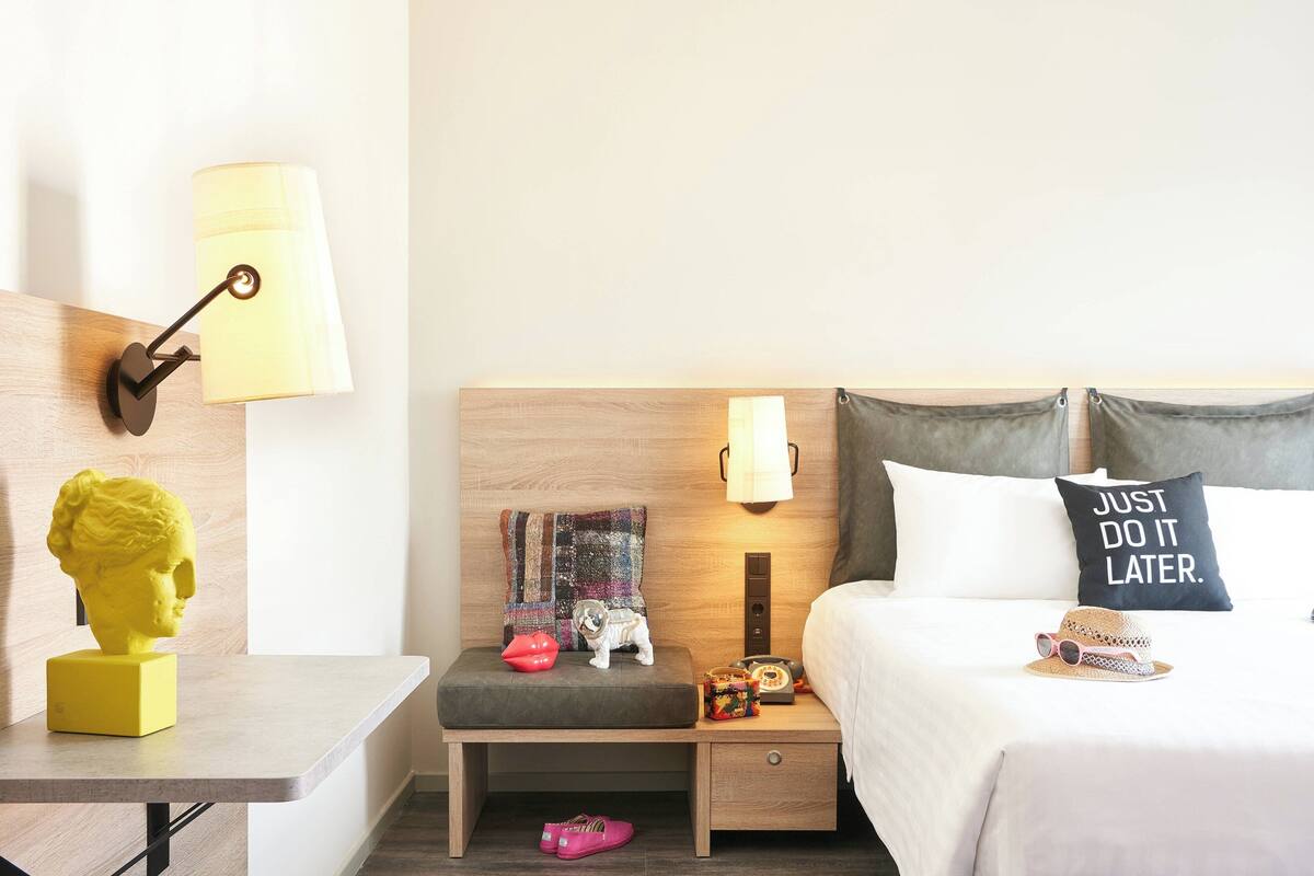 A hotel room with a wooden headboard, a bedside table with a lamp, a nightstand with a lamp, a bed with a pillow saying "JUST DO IT LATER.", a small bench with a blanket, a statue, and a hat.