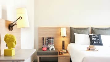 Quarto de hotel com cabeceira de madeira, mesa de cabeceira com abajur, um criado-mudo com luminária, cama com almofada que diz 'JUST DO IT LATER', pequeno banco com manta, estátua e chapéu.