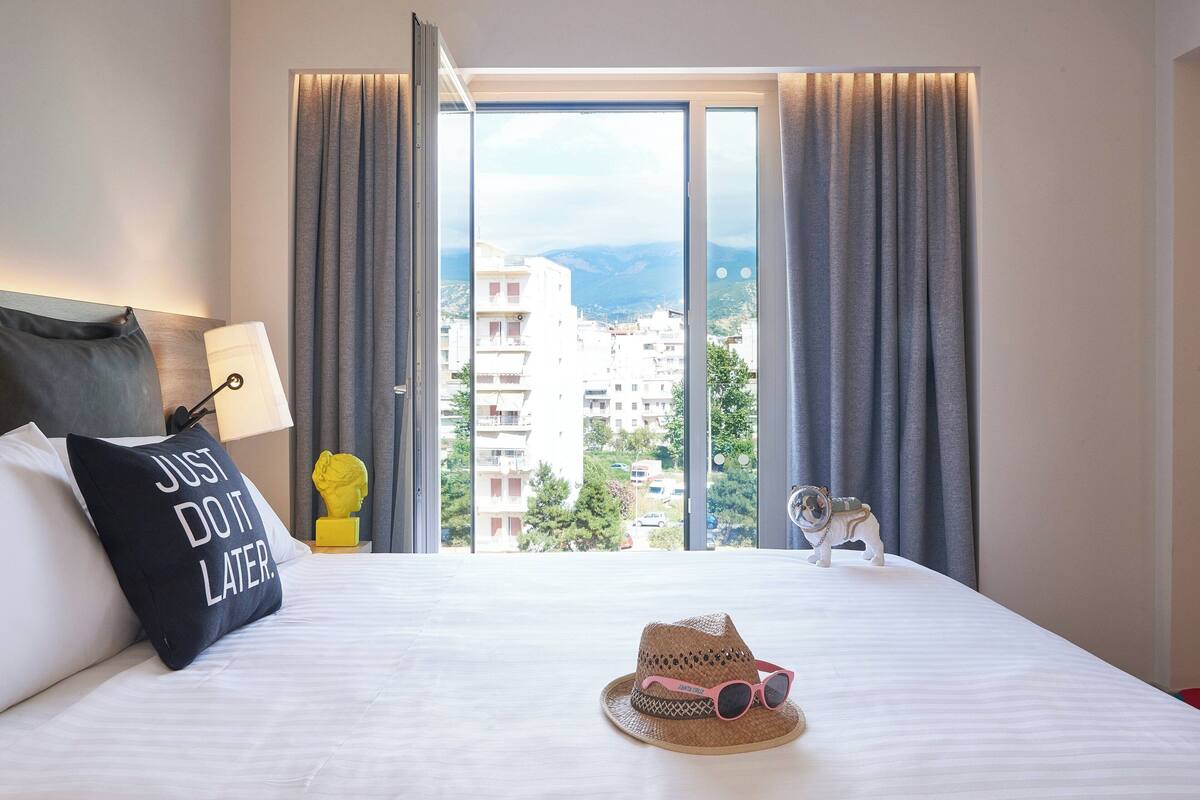 A hotel room with a large window, a bed with a pillow saying "JUST DO IT LATER", a hat, and a small figurine on the bed.