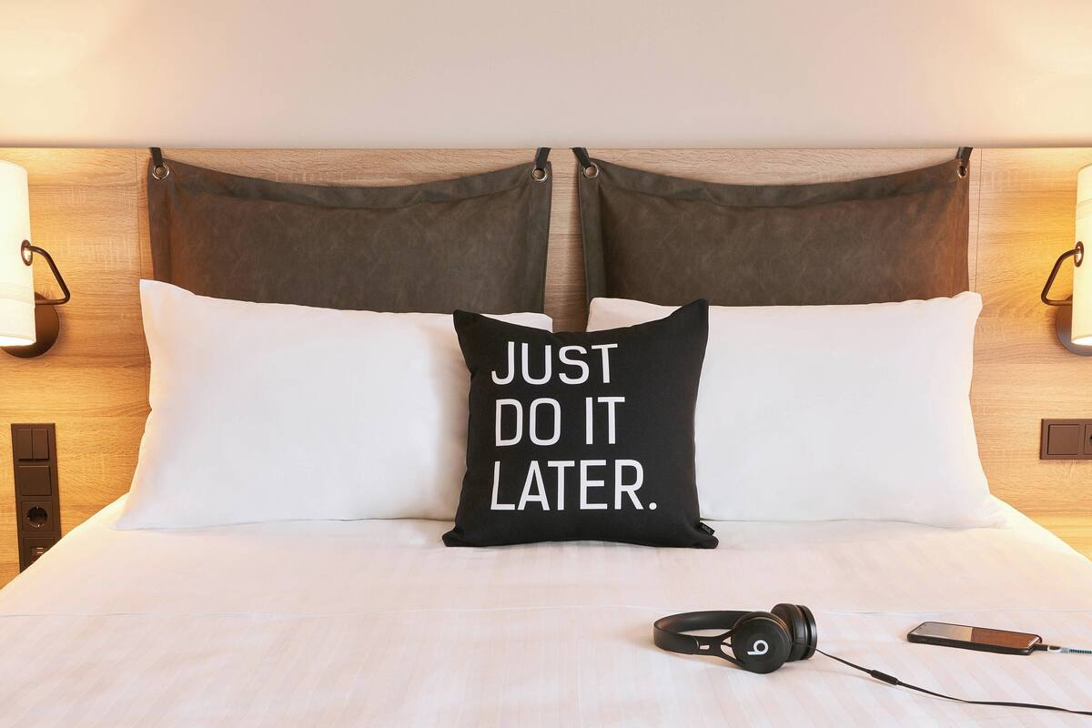 A neatly made bed with a black pillow featuring the text "JUST DO IT LATER." and a pair of headphones on the bed.