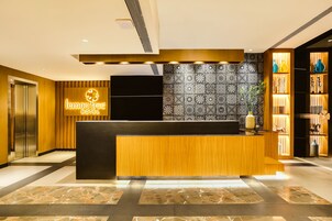 Zona de recepción de un hotel con un mostrador de madera, un papel tapiz decorativo y una estantería con objetos ornamentales.