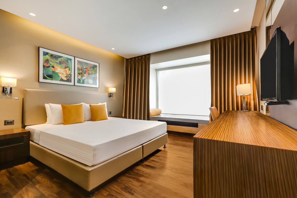 A modern hotel room with a large bed, bedside lamps, a wooden desk, and a large window with curtains.