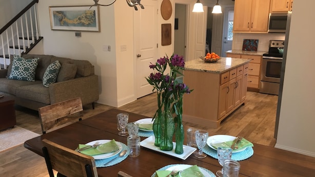 A dining area with a wooden table set for four, a vase with purple flowers, and a kitchen in the background. - PRICE REDUCED- includes linens and beach parking pass!  (Rehoboth Beach)
