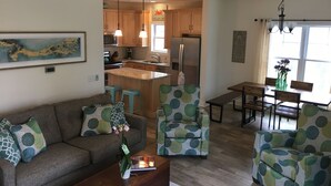 A modern living room with a sofa, two armchairs, a coffee table, and a dining area with a bench and table. A kitchen with wooden cabinets and stainless steel appliances is visible in the background. - PRICE REDUCED- includes linens and beach parking pass!  (Rehoboth Beach)