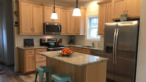A kitchen with wooden cabinets, a stainless steel refrigerator, a microwave, and a teal kettle on the stove. There are two blue bar stools and a bowl of oranges on the island. - PRICE REDUCED- includes linens and beach parking pass!  (Rehoboth Beach)
