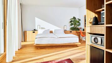Un dormitorio moderno con piso de madera, una cama con ropa blanca, un escritorio con planta y una cafetera integrada.
