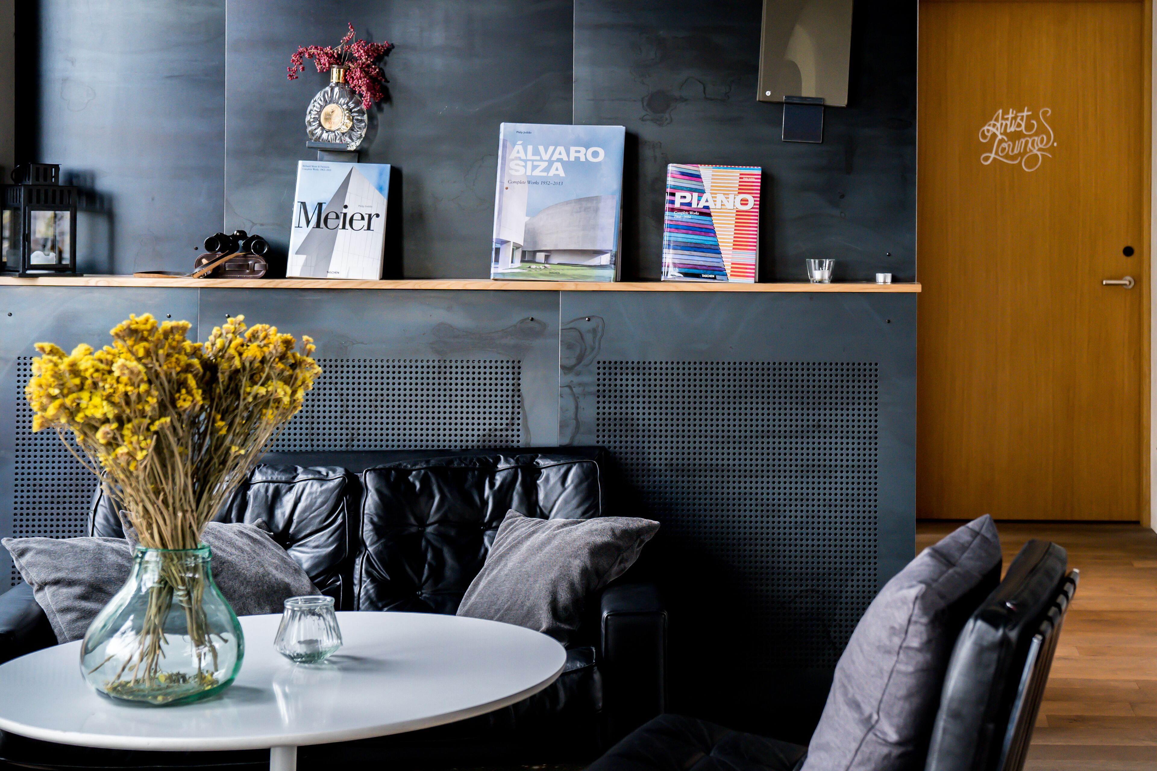 A modern reception area with a black leather couch, a round table with a vase of yellow flowers, and a wooden reception desk displaying books and a decorative clock.