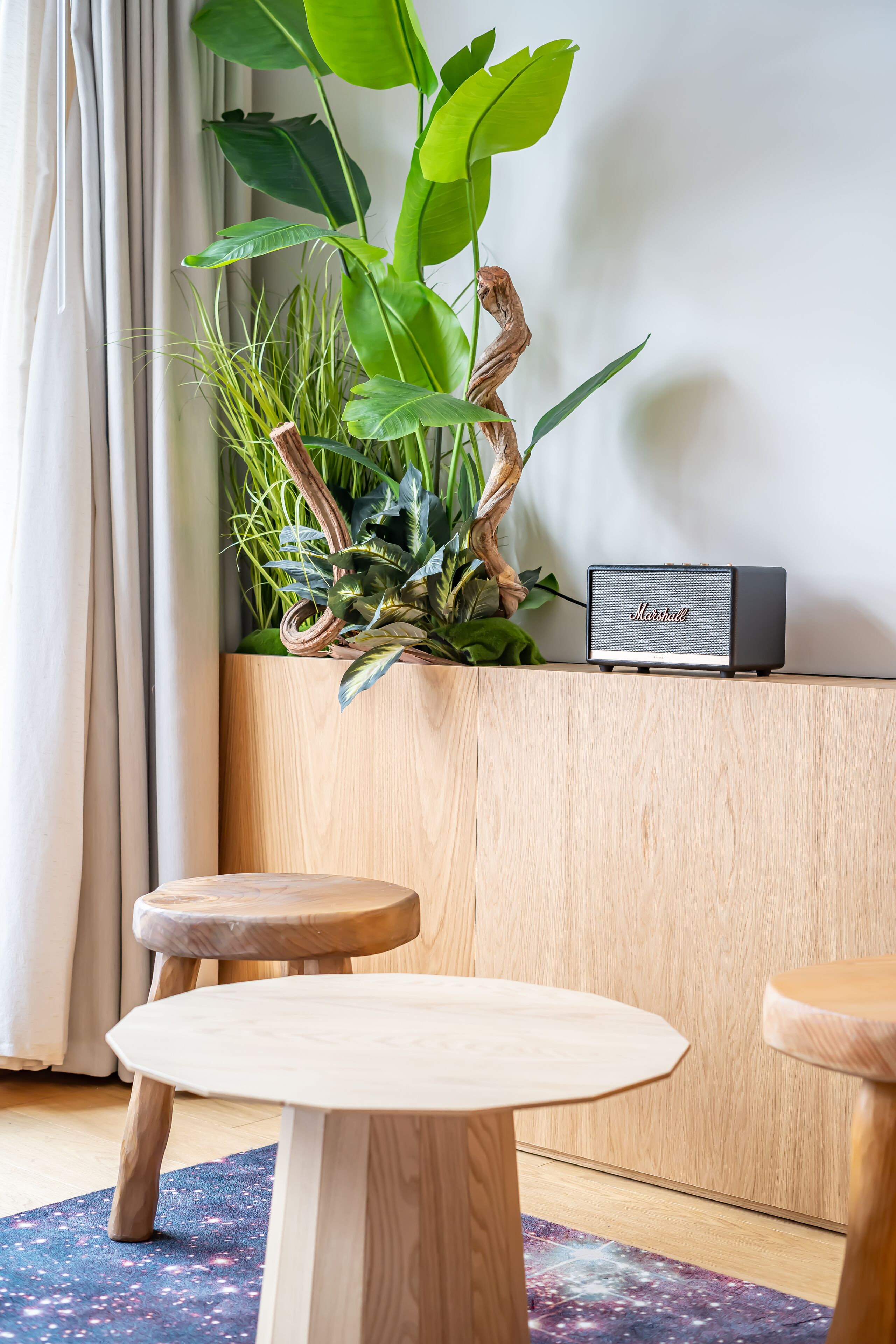 Una habitación con una mesa de madera, un taburete pequeño, una planta y un altavoz.
