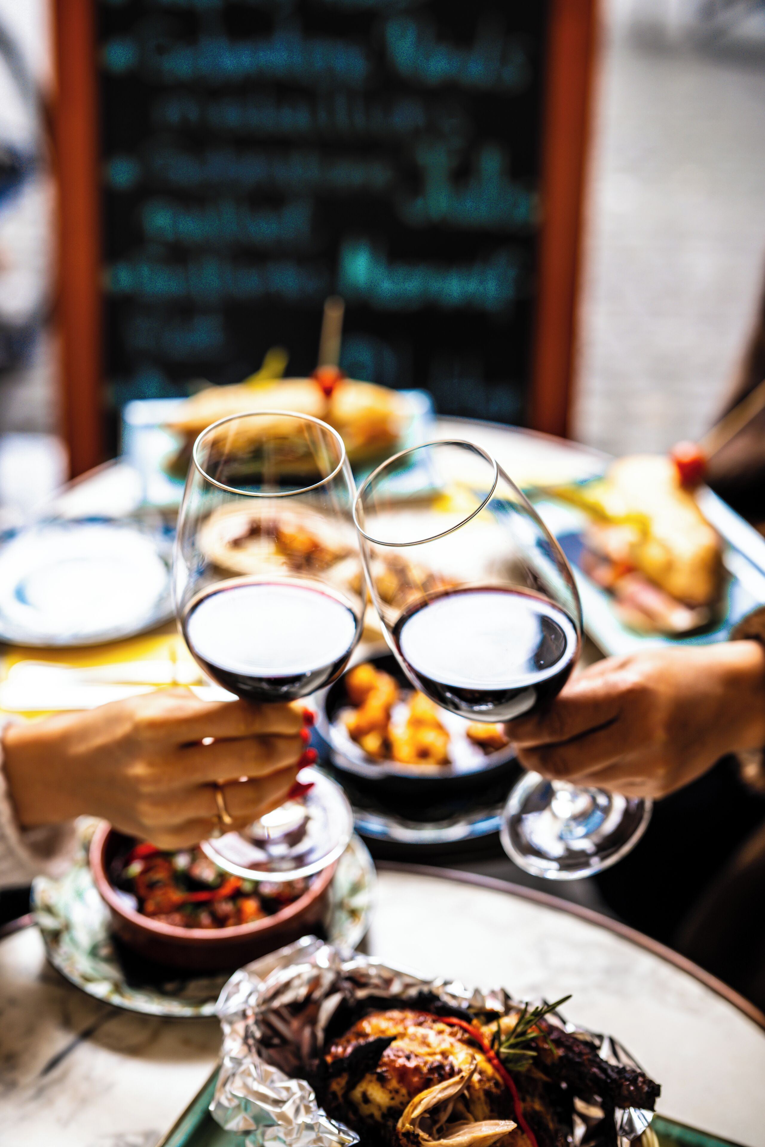 A dining table with plates of food, glasses of wine, and a chalkboard menu in the background.