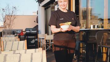Une personne tenant un bol, debout sur une terrasse aménagée avec des sièges et une table où sont disposés des aliments pour le petit-déjeuner.