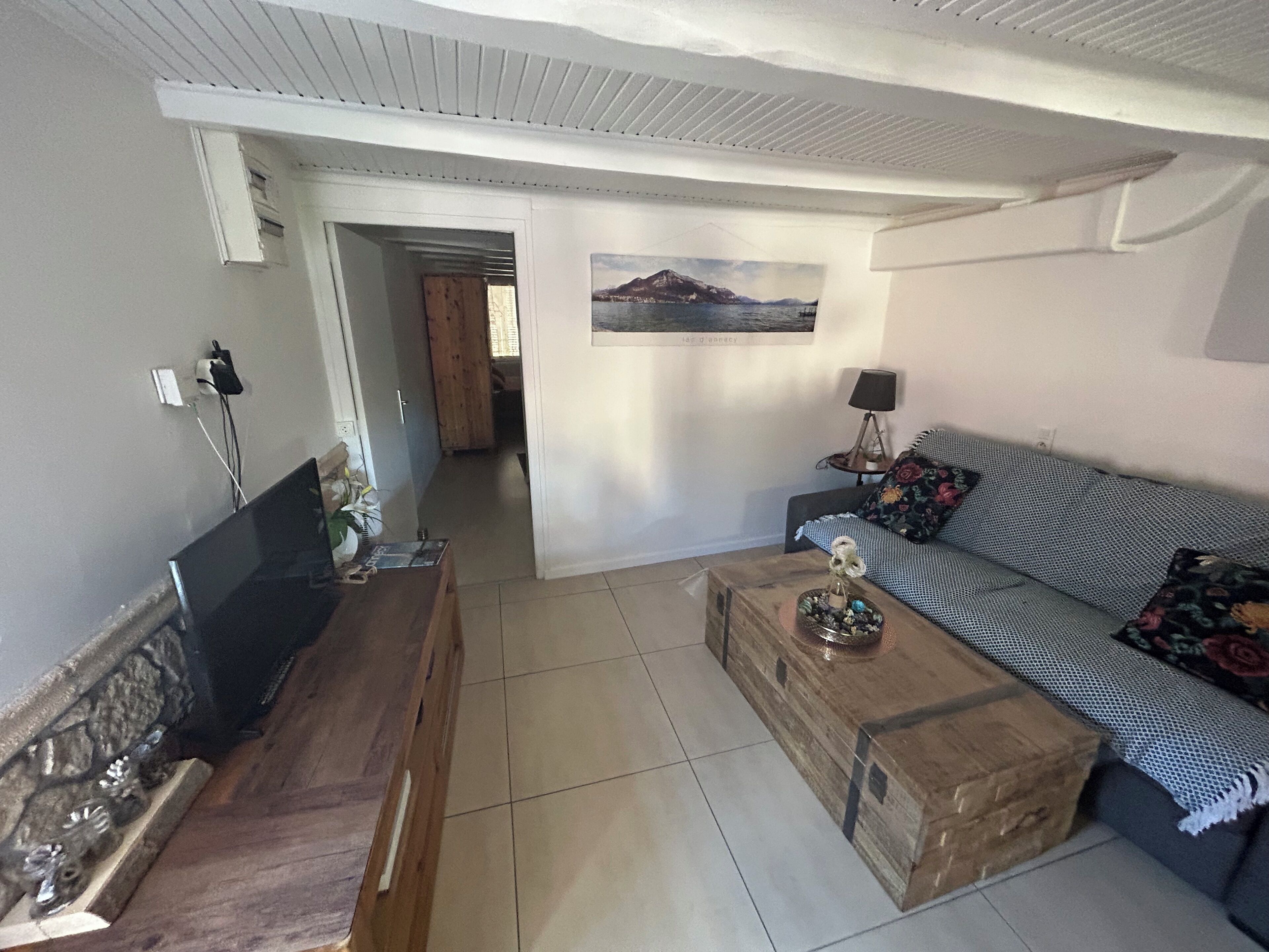 Un salón con una mesa de centro de madera, un sofá con cojines estampados, un cuadro en la pared y una puerta que da a otra habitación.