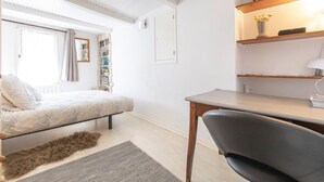 A small, well-lit bedroom with a bed, a desk, and a chair. There is a bookshelf filled with books and a bicycle hanging on the wall.