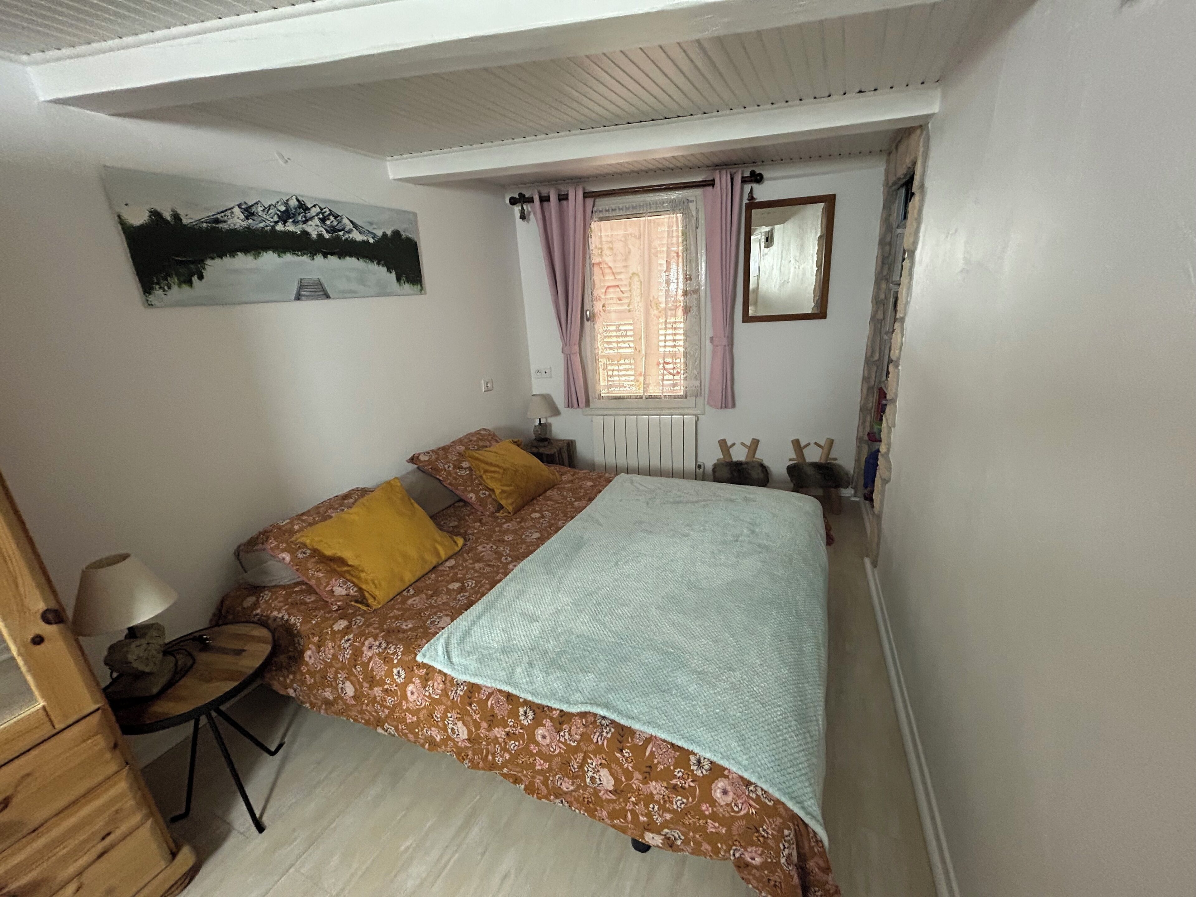 A bedroom with a bed, a desk, a chair, a window with curtains, a radiator, and a painting on the wall.