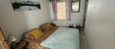 A bedroom with a bed, a desk, a chair, a window with curtains, a radiator, and a painting on the wall.