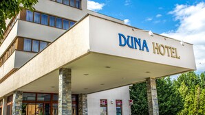 Il s’agit d’un hôtel à l’architecture moderne, qui se distingue par une enseigne bien visible portant l’inscription « DUNA HOTEL ». L’édifice compte plusieurs étages, de grandes fenêtres et une entrée abritée.