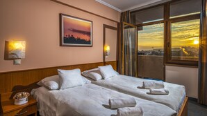 Une chambre d’hôtel avec deux lits, une vue sur la ville, un tableau représentant un coucher de soleil et une table de chevet ornée d’un vase de fleurs.