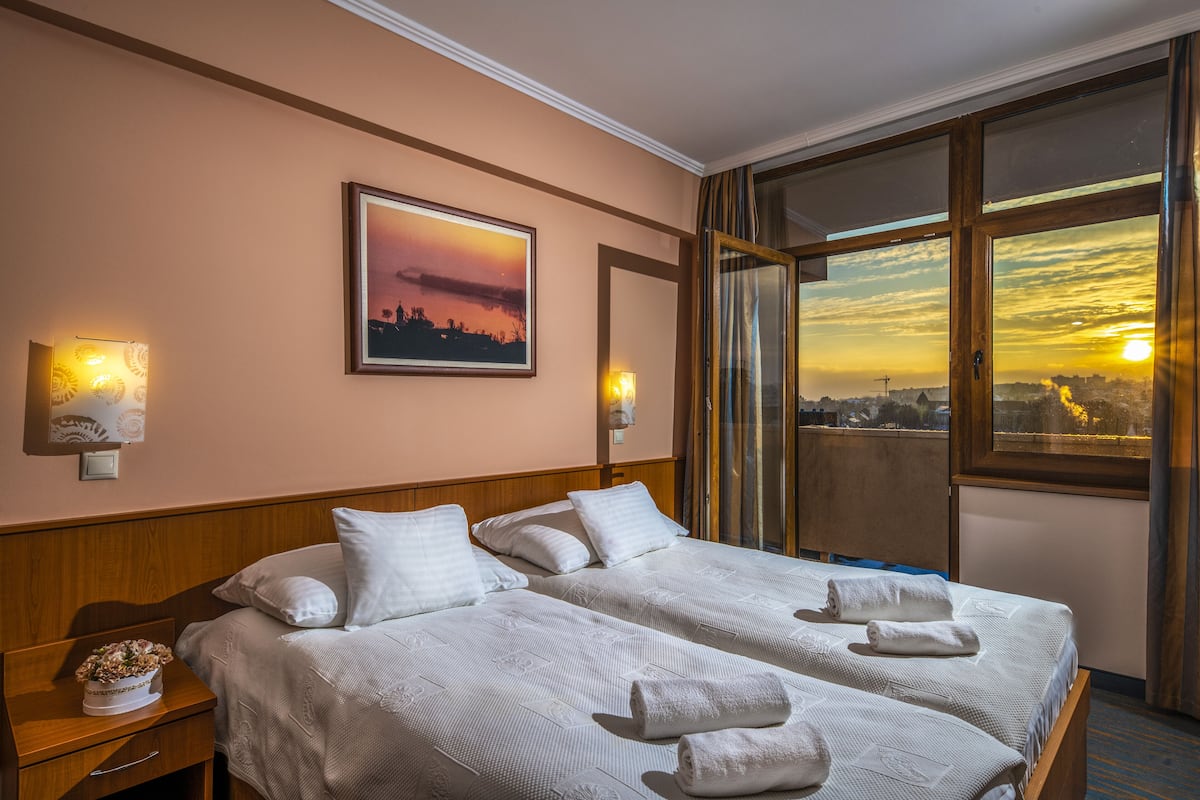 A hotel room with two beds, a city view, a sunset painting, and a nightstand with a flower vase.