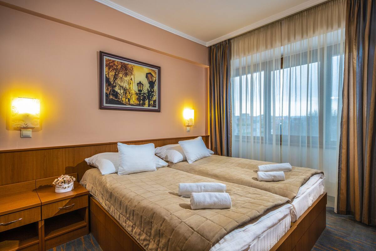 A hotel room with two beds, a nightstand, a framed picture on the wall, and a window with curtains.