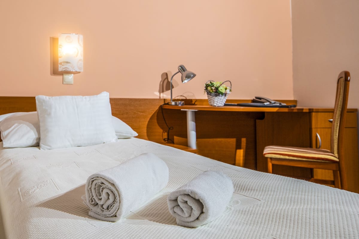 A neatly made bed with rolled towels, a desk with a lamp, a chair, and a wall-mounted light fixture.