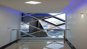 A hallway with a glass railing, a geometric ceiling design, and a view of buildings outside. - Crystal Inn (Chikkamagaluru)