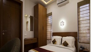 A hotel room with a bed, a desk, a chair, an air conditioning unit, and a window with blinds. - Crystal Inn (Chikkamagaluru)
