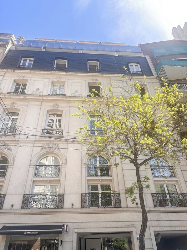 A multi-story building with balconies, a tree in the foreground, and a clear sky.