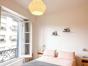 A bedroom with a bed, a window with curtains, a hanging light fixture, and a small bedside table with a plant.