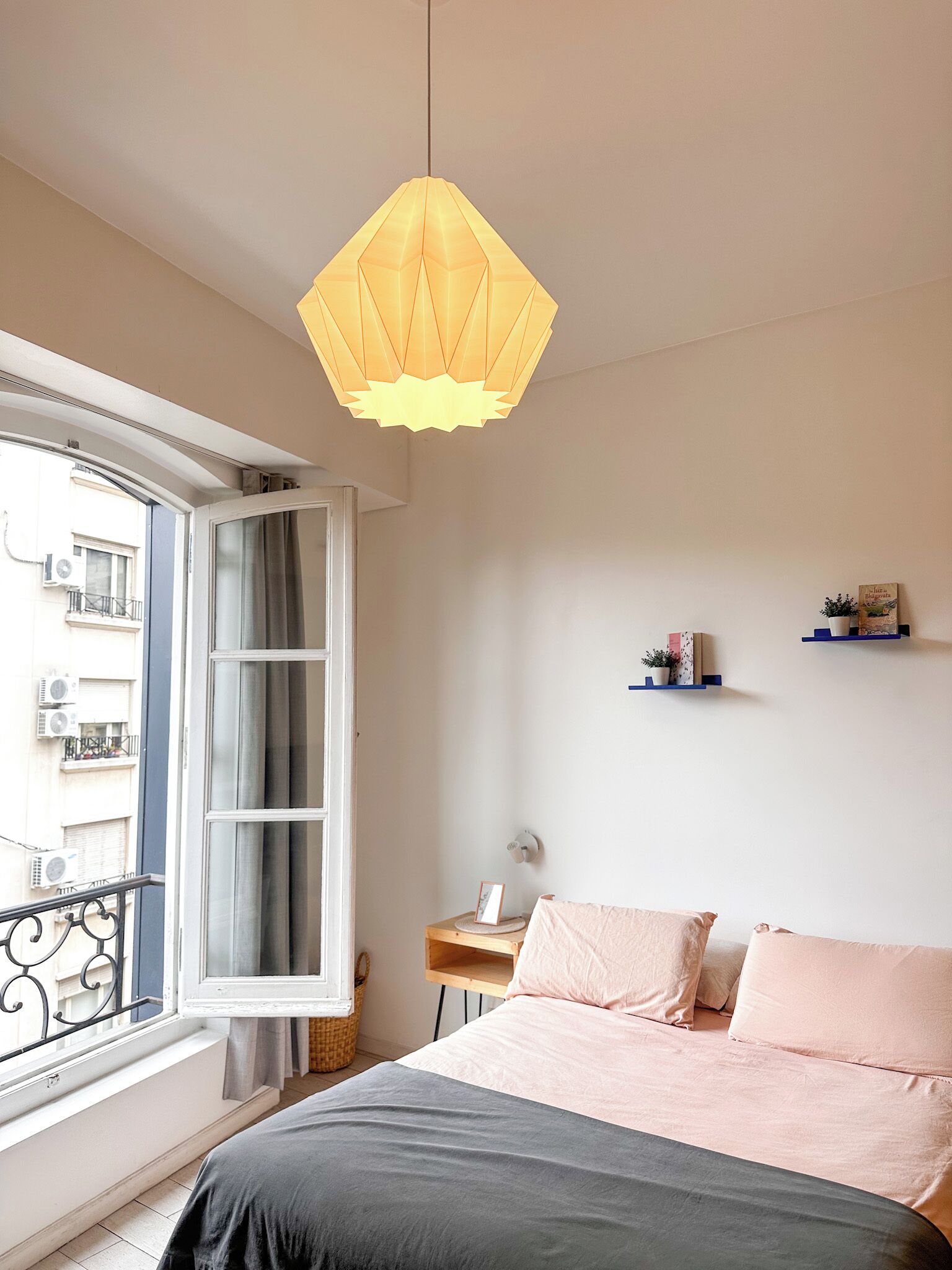 Un dormitorio con cama, ventana con cortinas, lámpara colgante, mesita de noche y estante en la pared.