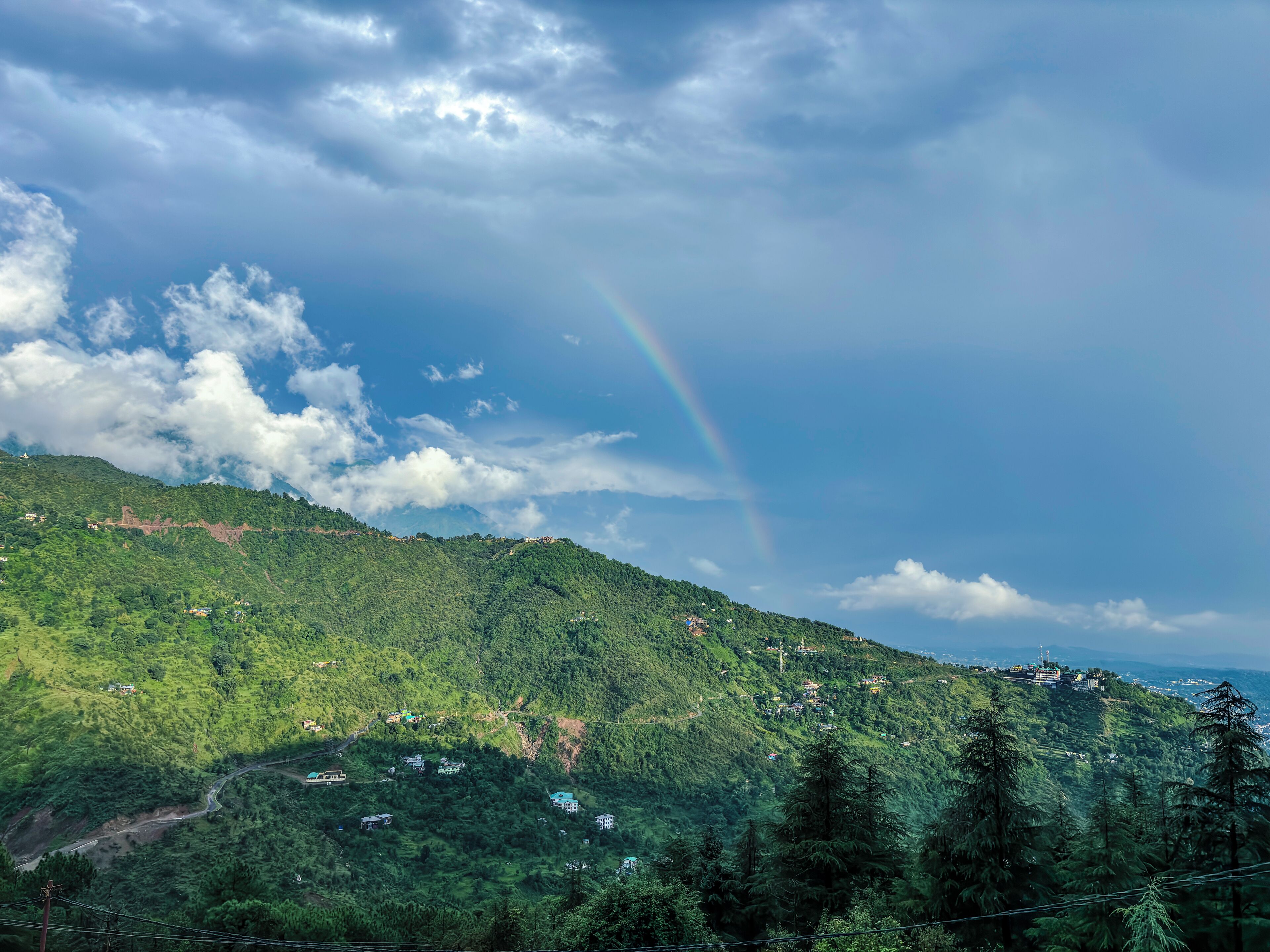 A lush green hillside with scattered buildings, a winding road, and a visible rainbow.