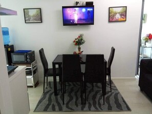 Un comedor con televisión montada en la pared, una mesa con sillas, microondas y una pequeña cocina.