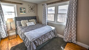 Quarto com cama, mesa de cabeceira, luminária, janela com cortinas e um objeto decorativo na parede.